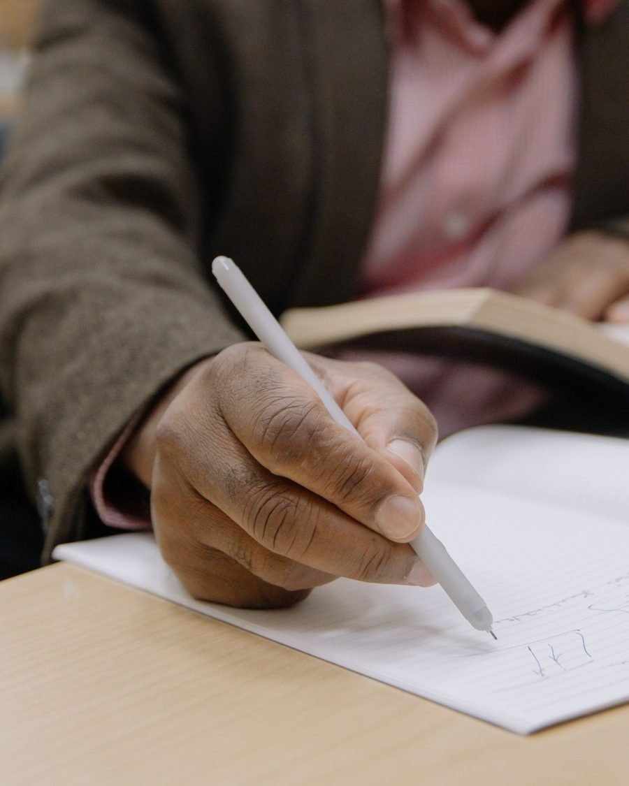An adult taking notes at a desk beside an open book